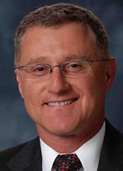 Headshot of a white man in a black suit with white shirt and glasses.