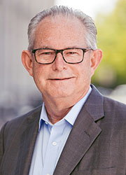 Headshot of a gray-haired white man in black glasses with a light-blue button down shirt and gray suit jacket.