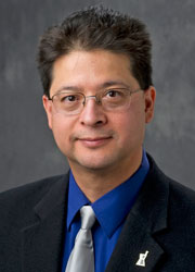 Headshot of Rodolfo Pinal, a man with short black hair wearing glasses, a blue shirt, silver tie, and black blazer