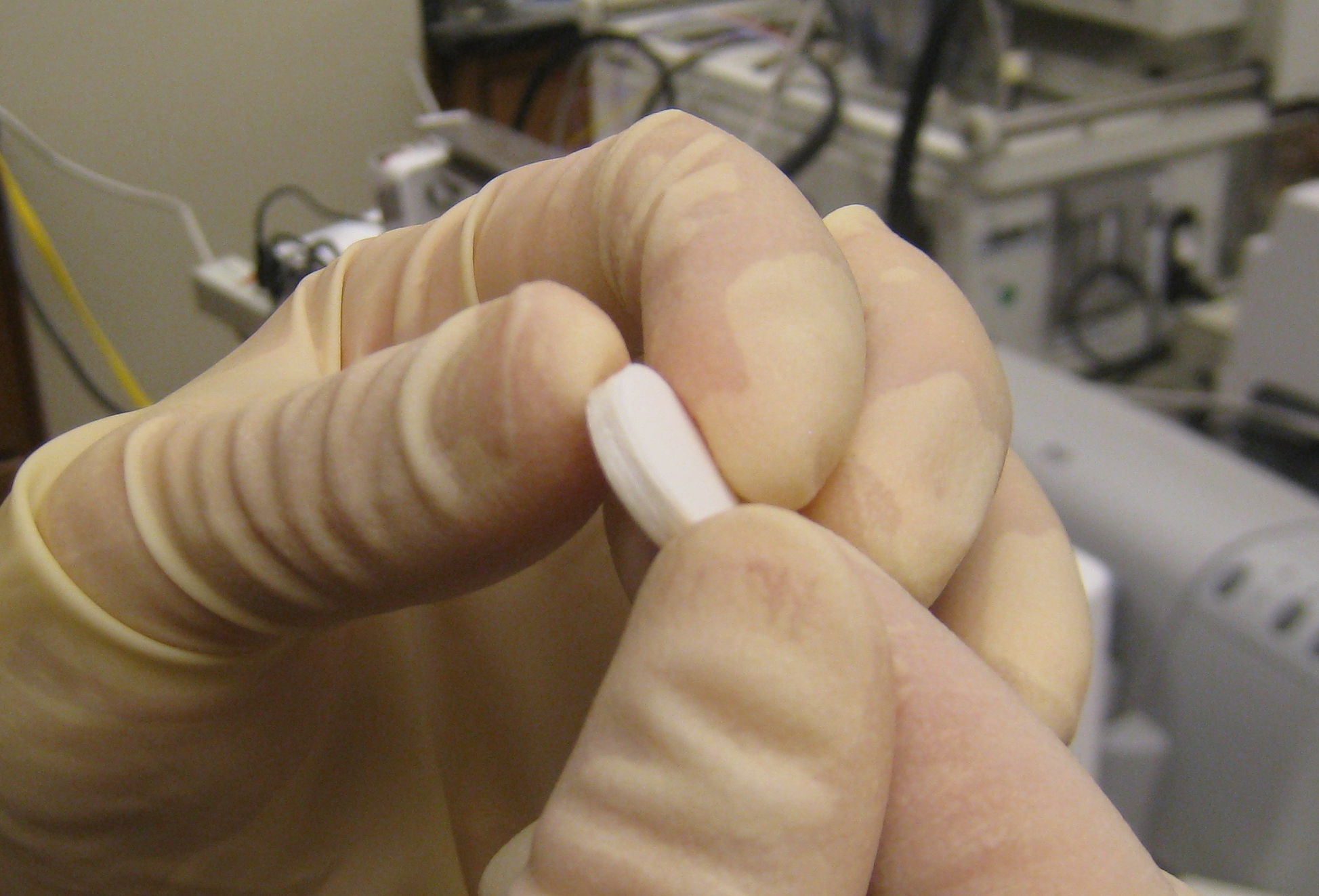 Fingers covered in off-white latex gloves hold a white pill with lab equipment in the blurred background.