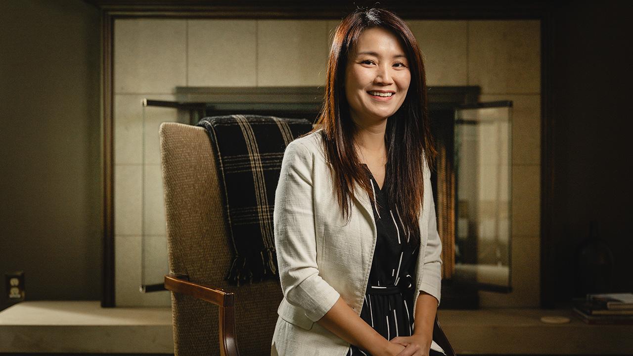 Purdue's First Lady, YingKei Hui, sits in a chair in front of a fireplace.