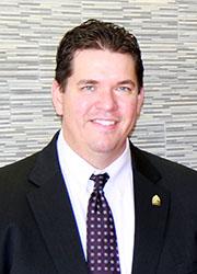 Headshot of a dark-haired white male in a black suit with patterned tie and white button-down.