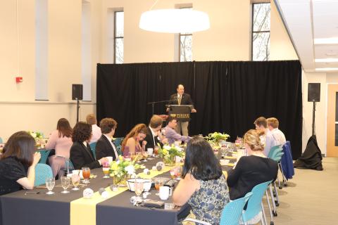 Graduating seniors sit at banquet tables while a male stands at a podium in front of a black backdrop addressing them.